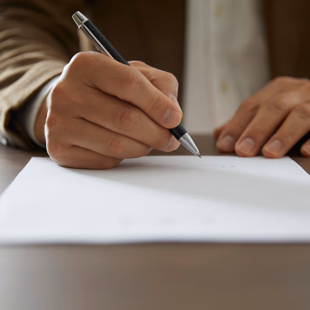 Close-up of a person holding a black pen poised above a blank white sheet of paper on a desk.